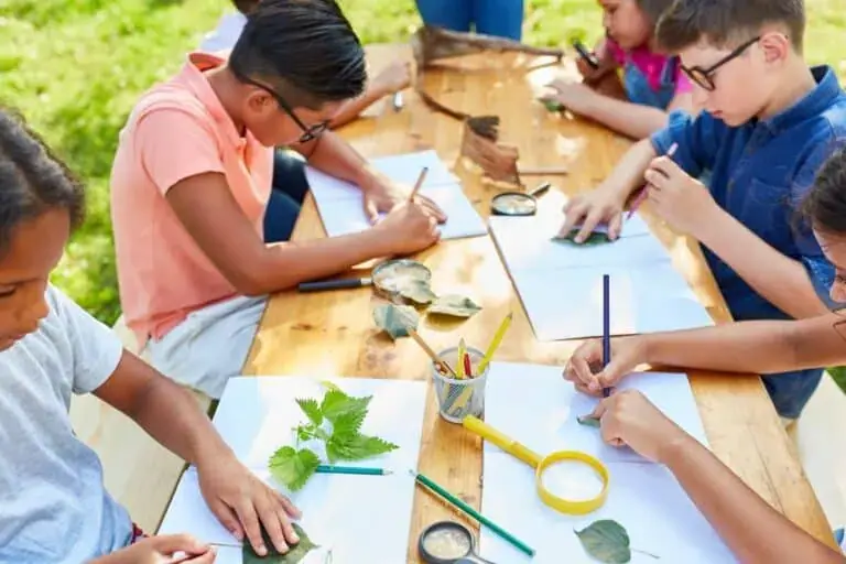 Children at an outdoor summer camp table working on nature science activities with leaves, magnifying glasses and notebooks